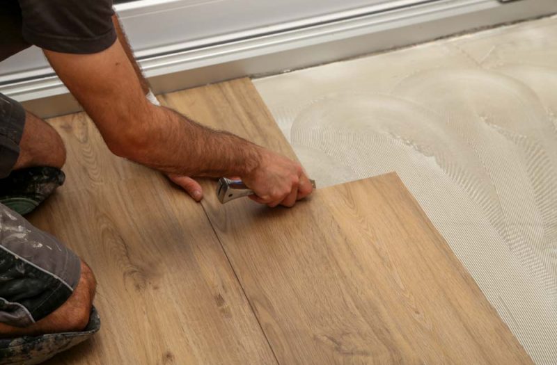 A detailed view of a worker trimming the edge of a wood laminate floor near a white doorway, using a utility knife for a precise fit.