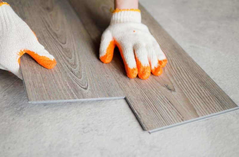 A professional laying dark grey laminate planks on a concrete floor, wearing orange safety gloves for protection and precision.